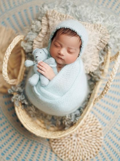 A cozy little cloud. This baby is nestled in a basket with a soft blue wrap and a tiny teddy bear, looking like he's floating on a cloud.
