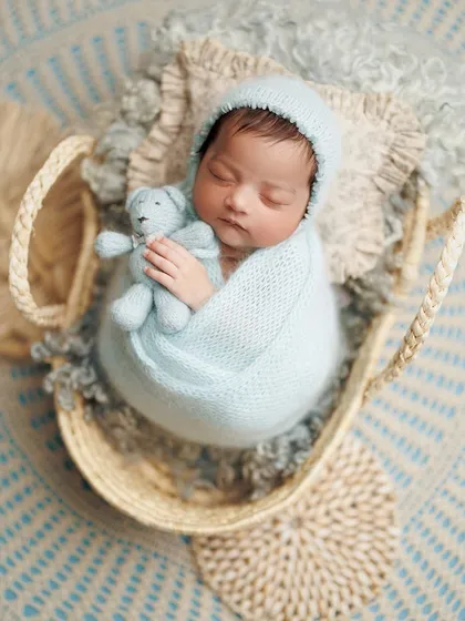A cozy little cloud. This baby is nestled in a basket with a soft blue wrap and a tiny teddy bear, looking like he's floating on a cloud.