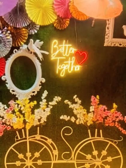 A trendy photo booth corner with a 'Better Together' neon sign on a green wall. The decorative bicycle and paper fans make it a fun spot for couple photos.