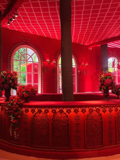 A custom-built bar for the 'Laal Ishq' event. The structure incorporates the theme's signature red color and intricate patterns, with large arched windows behind it offering a view of the gardens.