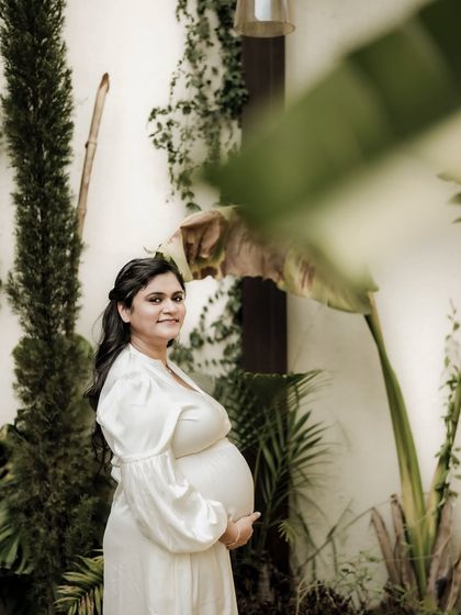 A beautiful portrait of the mom-to-be in a white dress, standing amidst lush greenery. The natural framing adds to the organic feel of the shot.