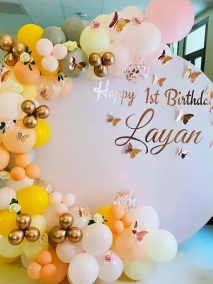 A 'Happy 1st Birthday' backdrop with a custom printed circular panel and a balloon garland in pastel yellow, orange, and grey.