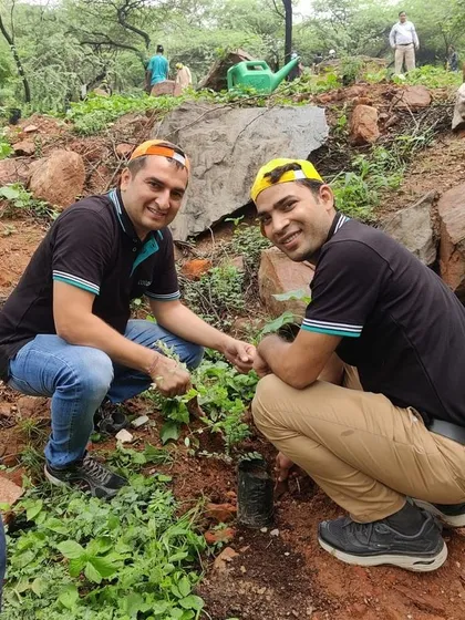 Two smiling volunteers from Colt Technology Services plant a sapling on a rocky incline at Aravali Nagar Van during a cloudy morning drive with 120 of their colleagues.
