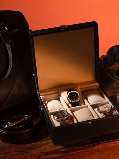 A lifestyle shot of a men's watch box, styled with other accessories like a belt and sunglasses. The warm, moody lighting creates a masculine and sophisticated atmosphere.