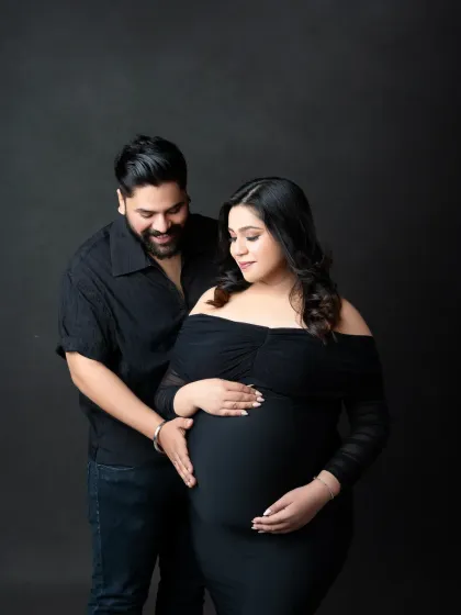 A classic and elegant maternity portrait against a solid dark grey backdrop. The couple is dressed in black, creating a dramatic and intimate look that focuses on their connection and the beautiful baby bump.
