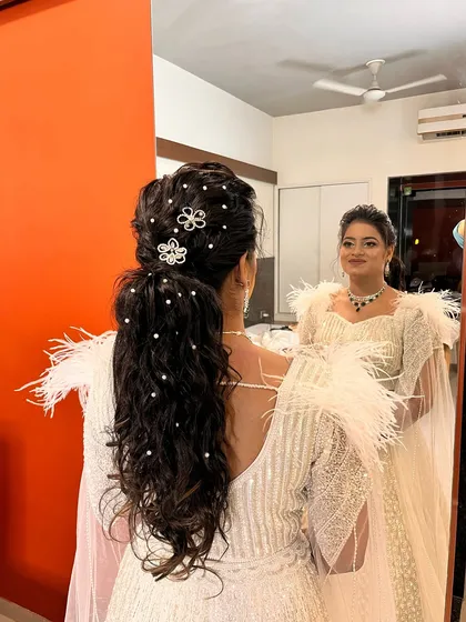 A crown of curls fit for a queen on her big day. This voluminous, pearl-studded ponytail is perfect for a reception or sangeet, paired with a glamorous white feathered outfit.