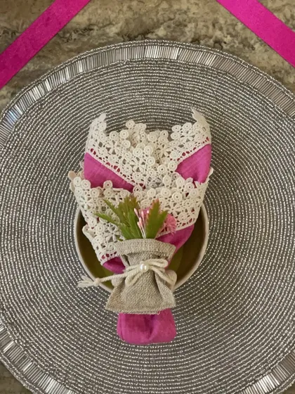 A close-up of a place setting from the pink ribbon theme. The napkin is folded into a delicate bouquet, combining a bright pink fabric with a lace doily and a small sprig of flowers for a touch of grace.