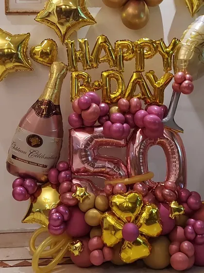 A festive balloon bouquet celebrating a 50th birthday. This arrangement uses pink and gold chrome balloons, large '50' foil numbers, and champagne bottle and glass foil balloons for a celebratory touch.