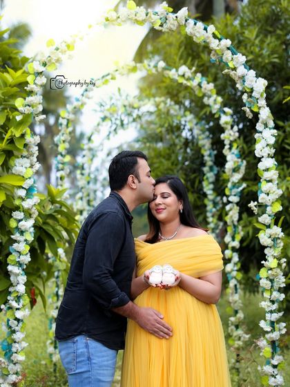A romantic couple's portrait under a floral archway, with the mother-to-be holding tiny baby shoes.