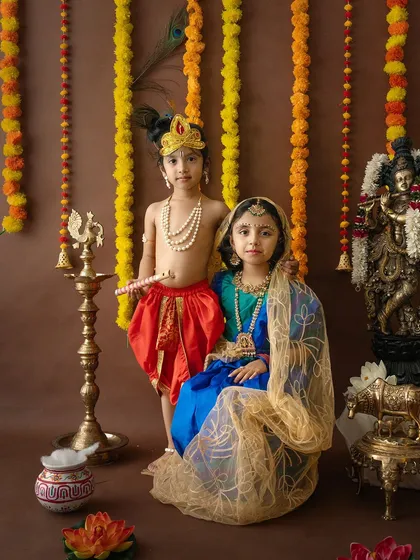 A lovely sibling portrait of a brother and sister dressed as Krishna and Radha, standing together in a complete festive setup.
