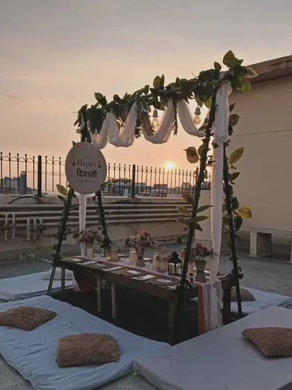 A beautiful sunset picnic setup on a terrace. The low table is set for a party, with a decorative sign and a frame adorned with greenery.