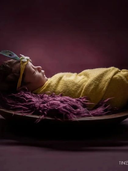 A simple yet striking portrait of a newborn with a peacock feather headband. The baby is swaddled in yellow and rests on a rich purple fur, creating a beautiful color contrast.