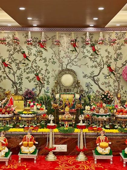A grand baby shower setup with a stunning backdrop of birds and trees. The foreground is filled with an elaborate arrangement of offering plates, dolls, and cow-and-calf figurines.