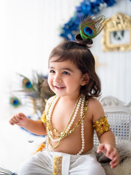 A close-up portrait of a happy little Krishna with a peacock feather in his hair bun.