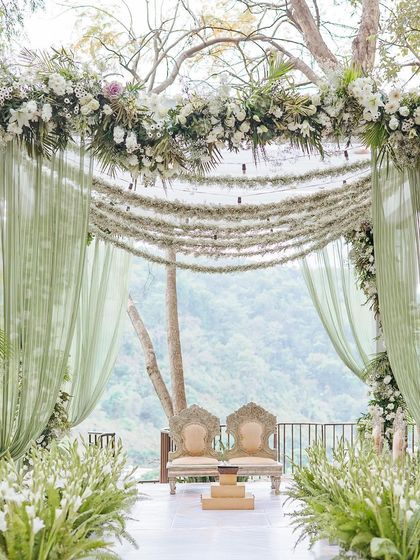 The stunning wedding mandap, with a canopy of white flowers and flowing sage green drapes, overlooking the forest.