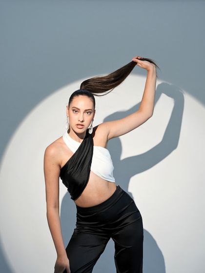 A striking black and white cutout top paired with black trousers. The high ponytail and spotlight effect create a dramatic, high-energy look for a modern fashion brand.