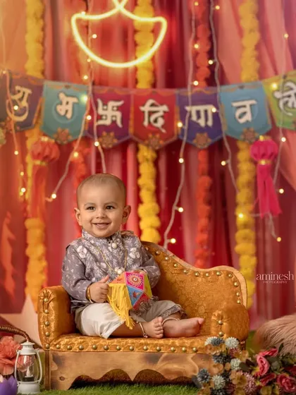 A smiling baby boy in a traditional kurta sits on a golden chaise lounge for his Diwali photoshoot. The background features a neon lamp and festive banners, creating a modern yet traditional feel.
