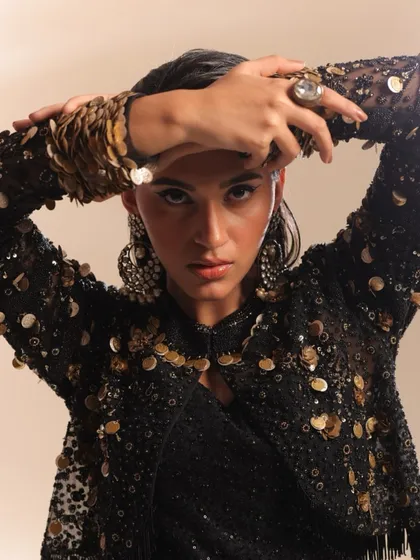 This close-up highlights a heavily embellished jacket with coin and sequin details. The model's pose, framing her face with her arms, draws attention to the intricate texture on the sleeves and the statement ring, creating a rich, tactile image.