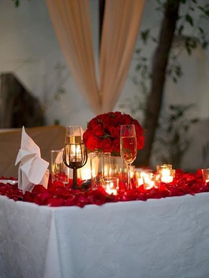 A close-up of a table prepared for a special night, with champagne flutes and a bouquet of red roses surrounded by candles and petals.