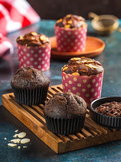 A collection of chocolate muffins, some in wrappers and some without, arranged on a wooden board to show texture and variety.