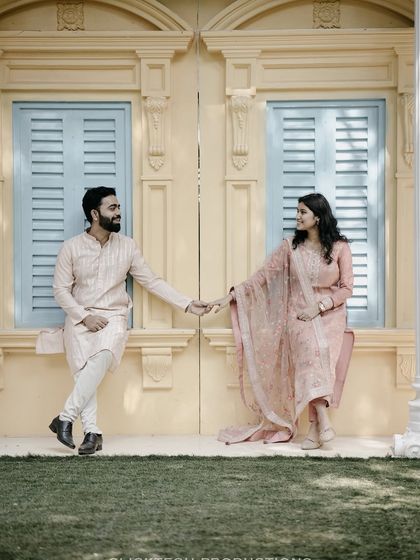 A symmetrically composed shot of a couple holding hands, posing against a colorful wall with blue-shuttered windows.