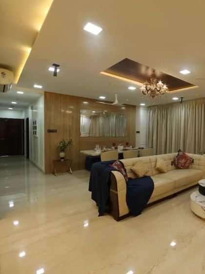 This spacious living room in a 3BHK at Oberoi Esquire, Goregaon, features an L-shaped sofa, marble-top center tables, and a grand chandelier, creating a luxurious and inviting atmosphere.