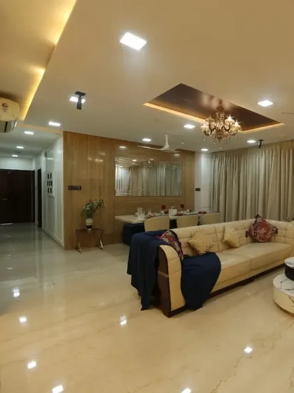 This spacious living room in a 3BHK at Oberoi Esquire, Goregaon, features an L-shaped sofa, marble-top center tables, and a grand chandelier, creating a luxurious and inviting atmosphere.