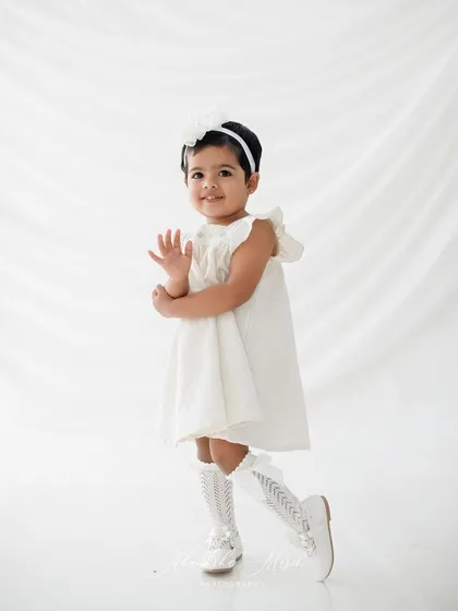 A playful wave from the birthday girl. Dressed in a classic white dress, she is all smiles and charm for her second birthday photoshoot.