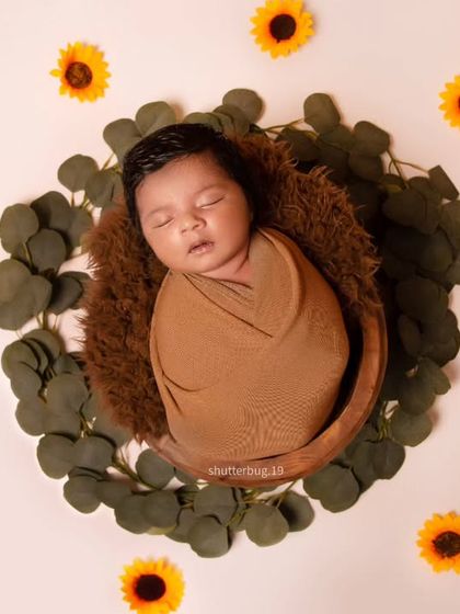 A classic swaddled newborn portrait taken from above. The baby is nestled in a basket lined with a brown fur blanket, set against a clean background with a wreath of leaves and sunflowers.