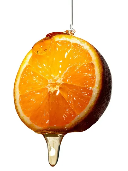 A slice of orange suspended and dripping with honey, shot against a clean white background. The hard light illuminates the fruit's segments and the viscosity of the honey, creating a beautiful abstract image.