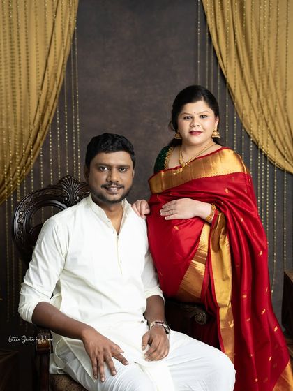A traditionally styled couple's portrait. The expecting mother, in a rich red silk saree, sits elegantly while her partner stands beside her in traditional white attire.