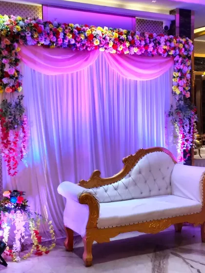 An elegant stage for a wedding reception or engagement, featuring a white and gold diwan, a backdrop of white and pink drapes, and a vibrant multi-color floral arch.