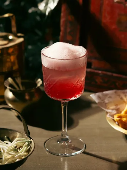 A beautiful pink cocktail with a frothy top. The use of traditional Indian brassware as props connects the modern drink to a classic aesthetic.