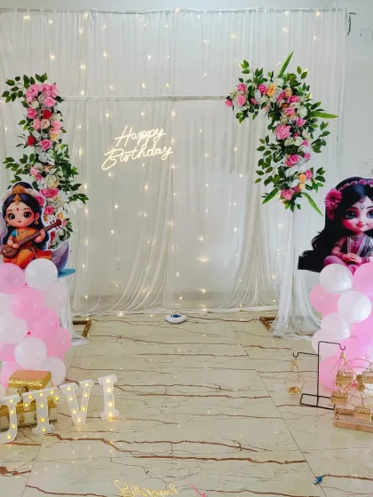 An elegant and serene birthday setup with a fairy light curtain backdrop. The decor is accented with floral arrangements and cutouts of Hindu deities, blending traditional and modern aesthetics.