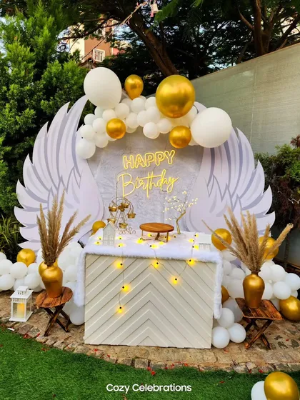 This elegant angel wing backdrop creates a heavenly photo opportunity for a special birthday. The design uses large white wings, a white and gold balloon garland, and a custom neon sign, perfect for an angelic or sophisticated party theme.