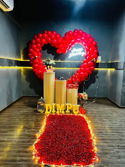 An indoor proposal setup featuring a large heart-shaped balloon arch made of red balloons. A neon sign, elegant pedestals for gifts or a cake, and a red petal carpet complete this romantic room decoration.