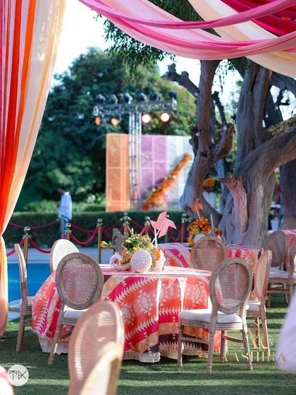 An outdoor dining table set under a canopy of colorful drapes, with a view of the pool and the stage in the background.
