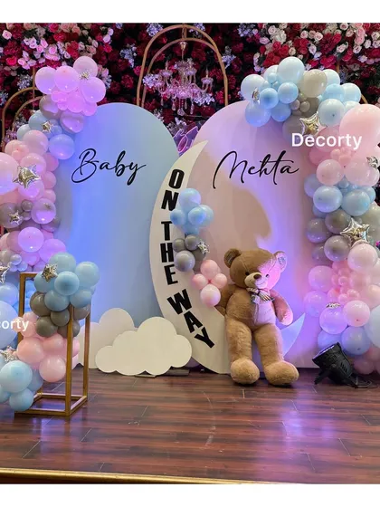 A chic and modern baby shower backdrop celebrating 'Baby Mehta on the way'. The design uses a soft blue and pink palette with a crescent moon detail, balloon garlands, and a large teddy bear for a dreamy welcome.