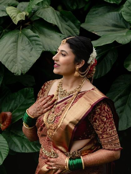 A candid shot of a bride, her hand to her chest. The rich maroon color of her saree and the intricate embroidery on her blouse are the focus of this beautiful image.