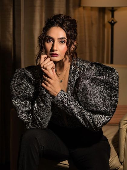 An indoor portrait of Ragini Dwivedi in a stylish silver and black top. The warm, soft lighting from the lamp creates an intimate and sophisticated mood.