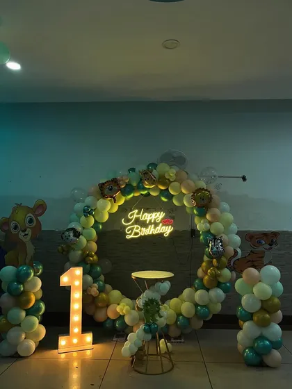 A circular balloon arch with a neon "Happy Birthday" sign forms the centerpiece for this jungle-themed party, flanked by character cutouts and a light-up number '1'.