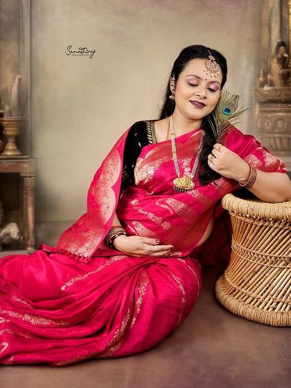 A serene portrait of a mother-to-be in a pink saree, holding a peacock feather. The classic props and rich background create a timeless, artistic feel.