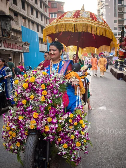 A woman rides a scooter adorned with a massive arrangement of flowers during the Gudi Padwa festival, with a traditional umbrella held behind her.