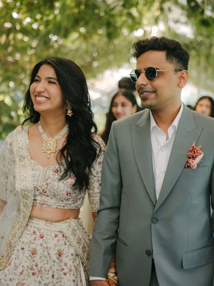 A candid shot of the happy couple walking through a garden path. The groom's modern sage green suit stands out, offering a fresh and sophisticated alternative to traditional wedding wear.