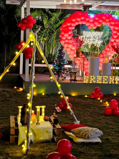A fun and festive birthday celebration combining two popular styles. A cozy, light-wrapped teepee for low seating is set in front of a large, red balloon heart backdrop with a "Happy Birthday" neon sign.