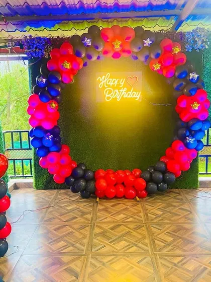 A bold birthday decoration with a circular balloon arch in red, blue, and black against a green wall, complete with a 'Happy Birthday' neon sign.