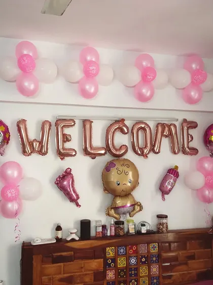 A simple and sweet 'Welcome' decoration for a baby girl. The setup uses pink and white balloons and various baby-themed foil balloons over a headboard.