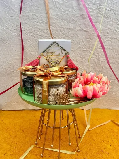 Celebrate the joy of Sankranti with this thoughtfully curated hamper on a green stand. It features jars of traditional sweets like til-gud, making it a perfect gift for the harvest festival.