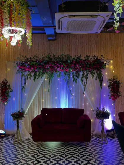 This is an elegant stage setup I created for a Roka ceremony in a restaurant. The combination of a rich red velvet sofa, a lush floral arrangement overhead, and soft fairy lights against a white curtain backdrop creates a sophisticated and romantic ambiance.