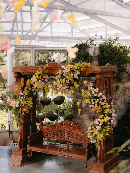 A traditional wooden swing, or 'jhoola', beautifully decorated for the Haldi ceremony. It's adorned with cascading arrangements of yellow and pink flowers, creating a perfect focal point within the greenhouse.